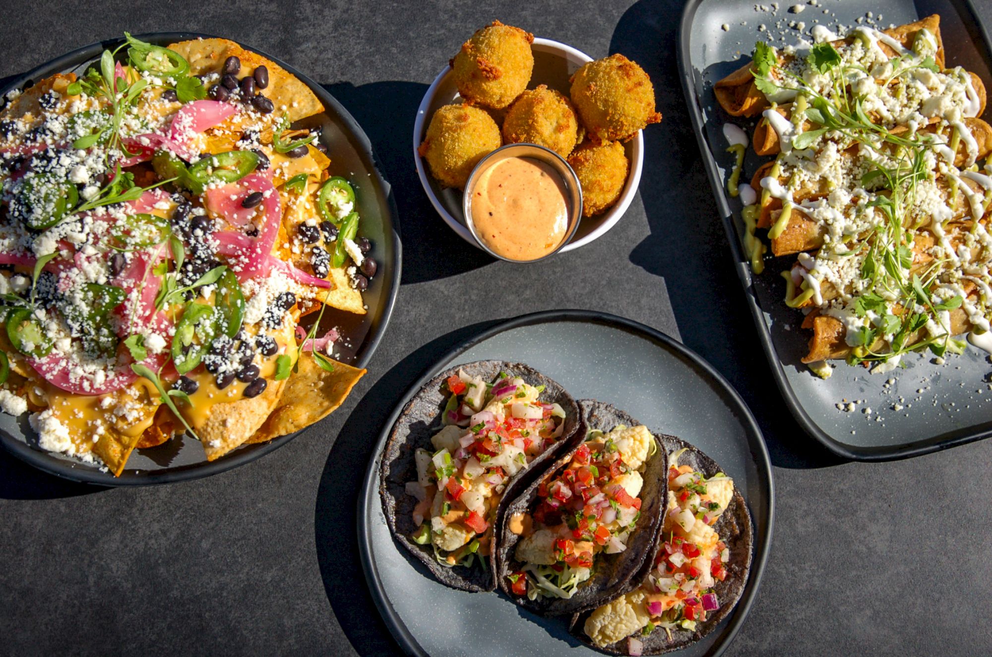 The image shows a spread of Mexican dishes, including nachos, tacos, croquettes, and a plate of loaded fries with dipping sauce in the center.