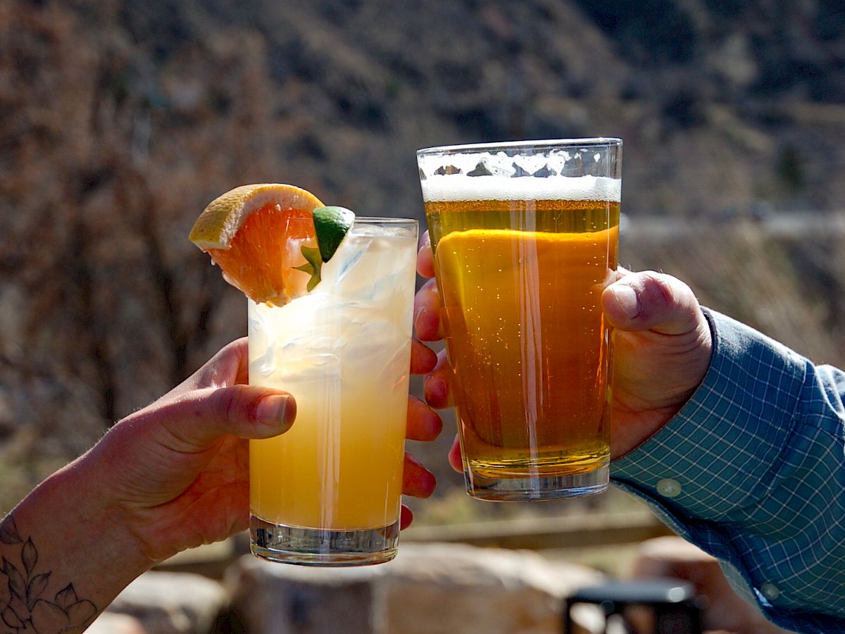 Two people clink glasses outdoors, one with a cocktail garnished with lime and the other with a pint of beer, against a mountainous background.