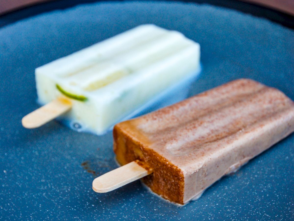 The image shows two popsicles, one white and the other brown, placed on a dark blue plate.