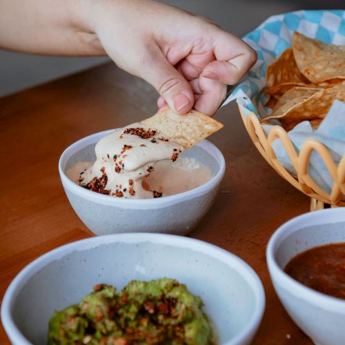 A hand dips a chip into a bowl of dip with visible toppings; nearby are bowls of guacamole and salsa, and a basket of tortilla chips.