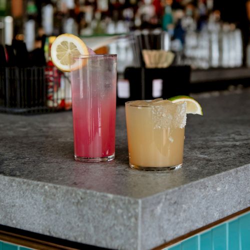 Two cocktails on a bar counter: a tall pink drink garnished with lemon and a short yellowish drink garnished with a lime slice.