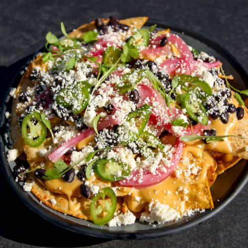 A plate of loaded nachos topped with cheese, jalapeños, pickled onions, black beans, and assorted fresh greens is shown.