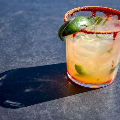 A cocktail in a glass with a spicy rim, garnished with lime slices, sits on a dark surface, casting a shadow.