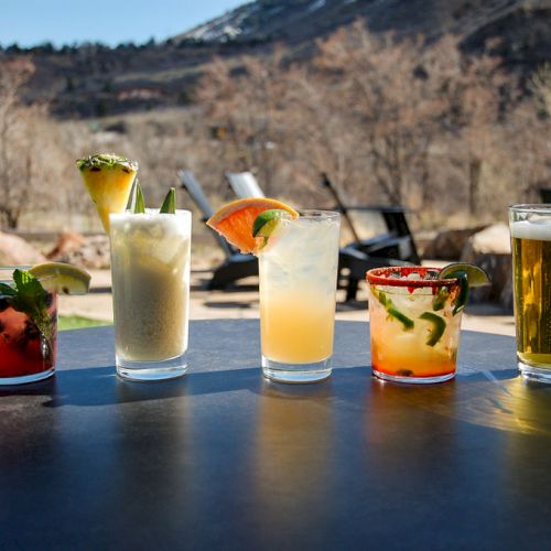 A variety of colorful drinks, including cocktails and a glass of beer, are set on a table with an outdoor mountain view in the background.