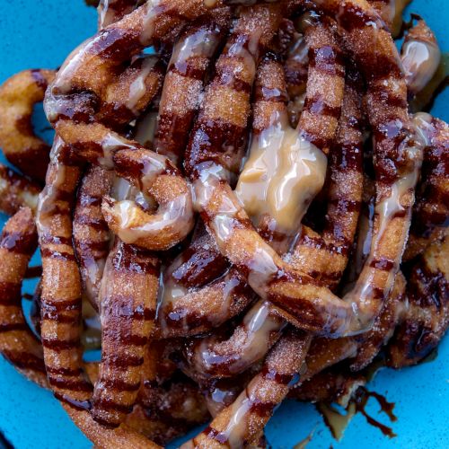 The image shows a plate of churros drizzled with caramel sauce on a blue plate, which are sweet, fried dough pastry sticks commonly enjoyed as a dessert.