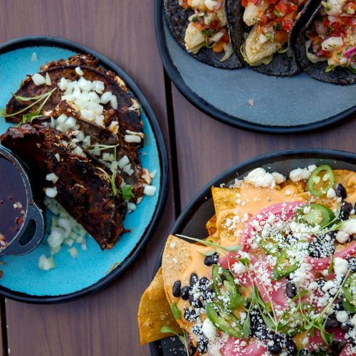 The image shows three plates of food: tacos on two plates and nachos on the third plate, all placed on a wooden table.