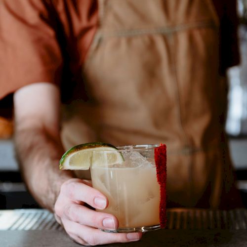 A person in an apron holds a cocktail garnished with a lime slice, standing by a counter in what appears to be a bar or café setting.