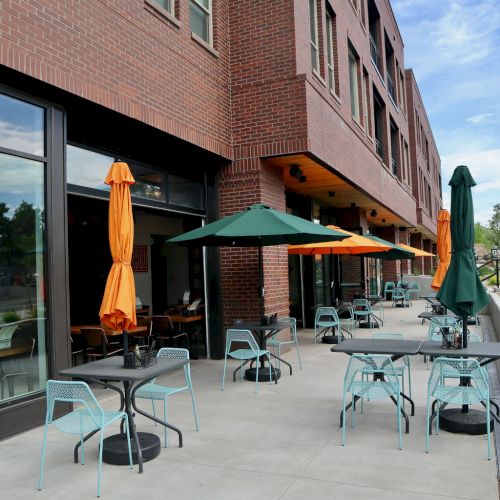 An outdoor seating area with tables, chairs, and umbrellas beside a brick building under a partly cloudy sky, shown in the image.