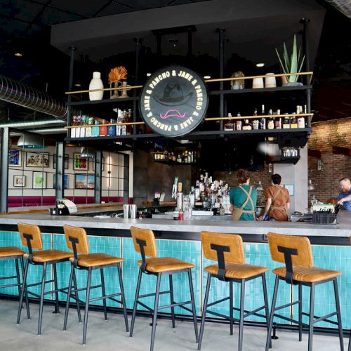 The image shows an indoor bar setup with stools, a counter, bartenders working, and shelves with various items and decor on display.