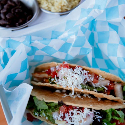 The image shows two tacos filled with fresh ingredients, served on a blue checkered paper, with sides of black beans and rice in small bowls.