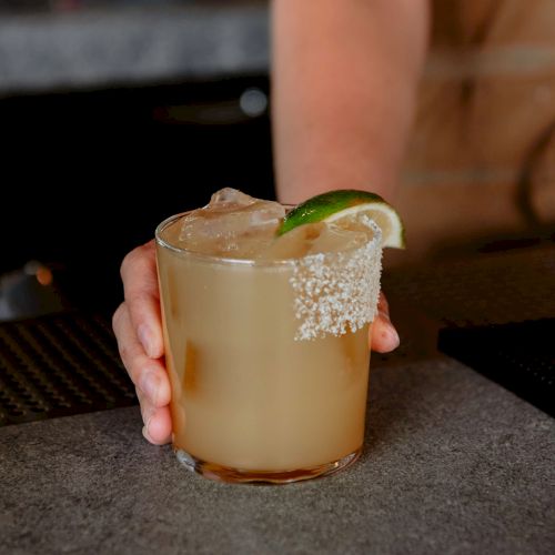 Someone holding a cocktail garnished with a lime wedge, with salt around the rim, likely a margarita, set on a bar counter.