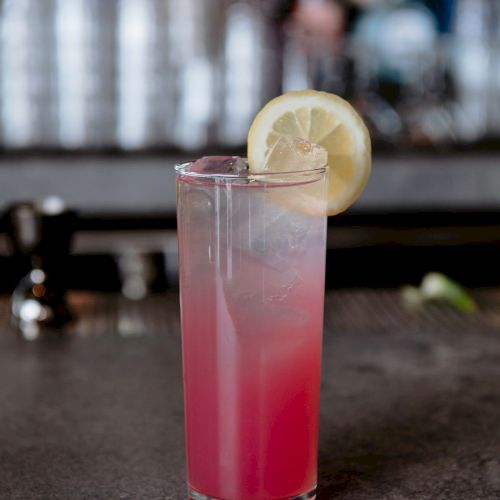 A tall glass filled with a pink-hued drink, garnished with a lemon slice, placed on a bar counter with blurred bottles in the background.