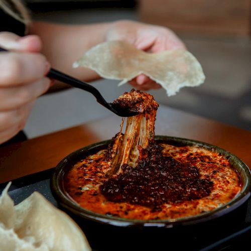 A person is using a fork to eat a cheesy, seasoned dish from a bowl, with bread on the side, creating a visually appetizing scene.