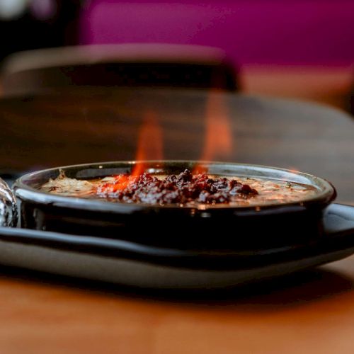 A dish on a tray with visible flames, possibly a dessert or flaming entrée, with a foil-wrapped item beside it on a wooden table.