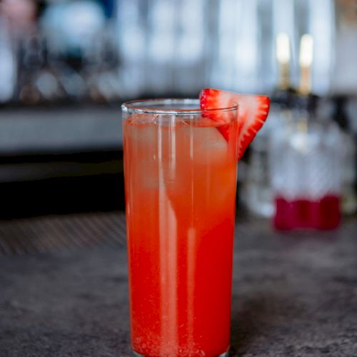 The image shows a tall glass filled with a reddish-orange drink, garnished with a slice of strawberry. The background is a bar setting.