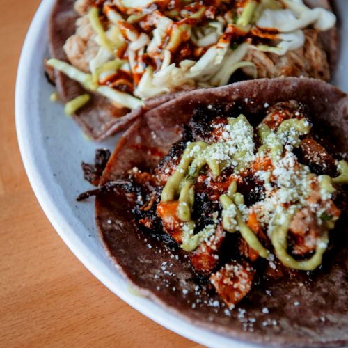 The image shows two tacos on a plate, garnished with avocado sauce, shredded cabbage, and various other toppings, displayed on a wooden table.