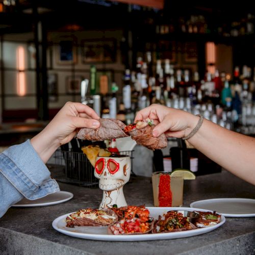 Two people are toasting with drinks over a plate of food in a bar with a TV and bottles in the background.