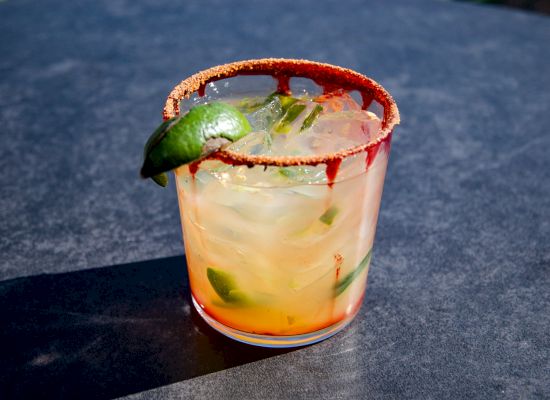 A refreshing cocktail with ice, lime, and a chili-powder-rimmed glass, placed on a dark surface.
