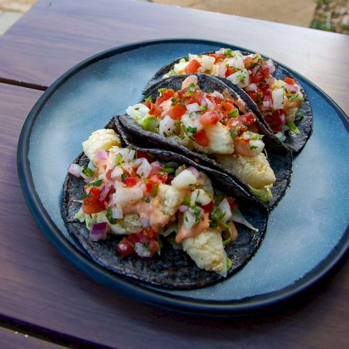 The image shows three tacos on a blue plate. The tacos have dark-colored tortillas, scrambled eggs, diced tomatoes, onions, and cilantro.
