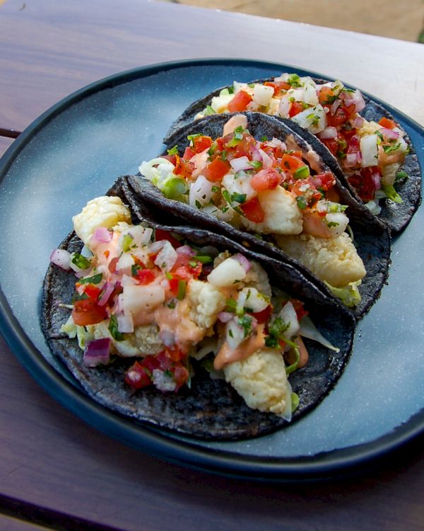 The image shows three tacos on a blue plate. The tacos have dark-colored tortillas, scrambled eggs, diced tomatoes, onions, and cilantro.