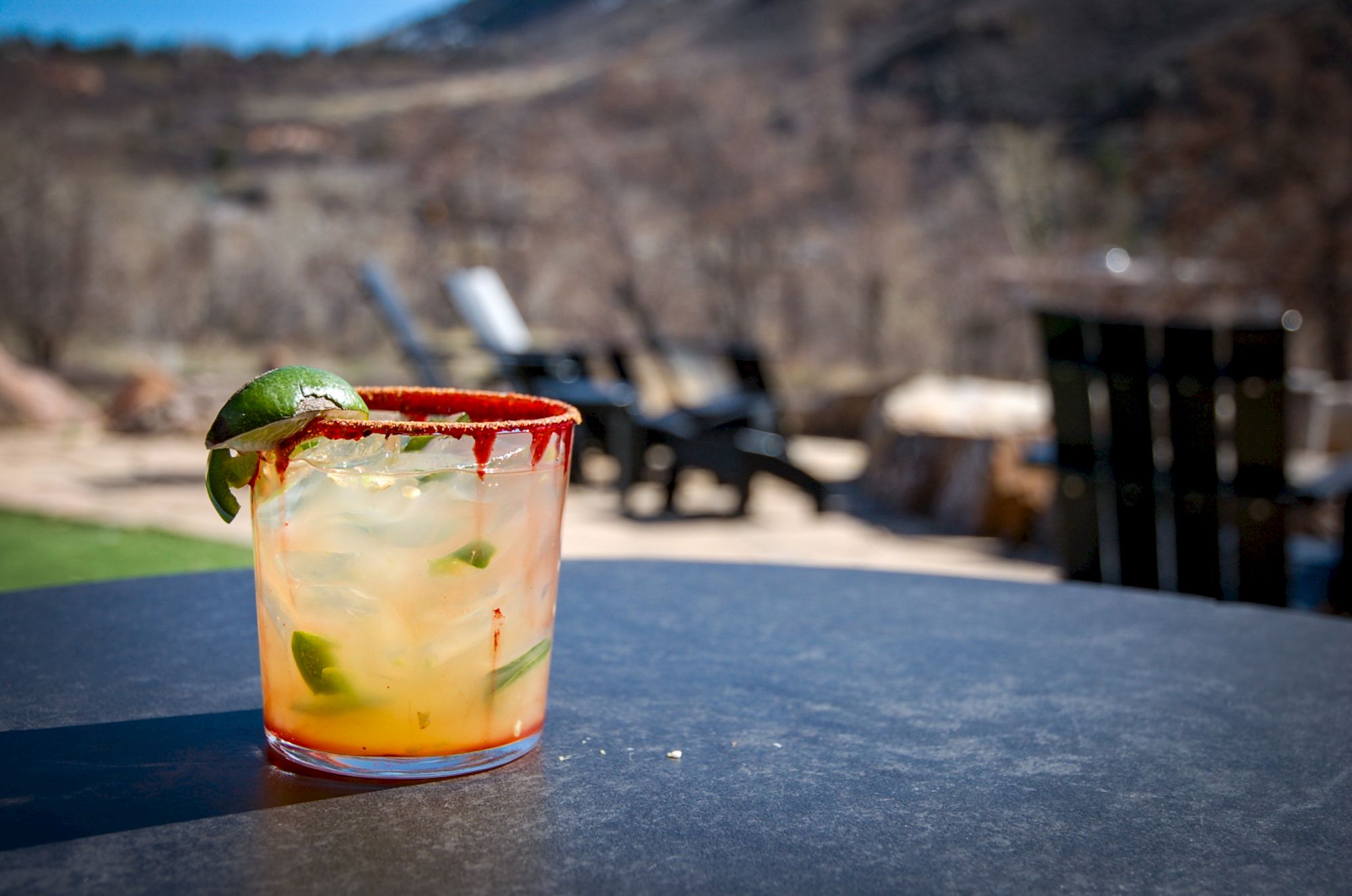 A cocktail garnished with lime and a chili-rimmed glass sits on a table outside with an out-of-focus scenic background and empty chairs.