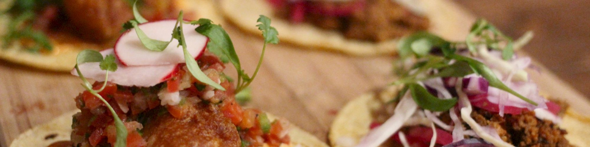 The image shows a wooden platter with four types of tacos, each topped with various ingredients like meat, vegetables, and garnishes.